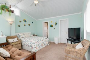 A bedroom with a bed, two wicker chairs, a television, and decorative wall art. - Blackbird Inn: Oceanside, Pet-friendly, Close to Beach, Short Stays Anytime! (Kill Devil Hills)