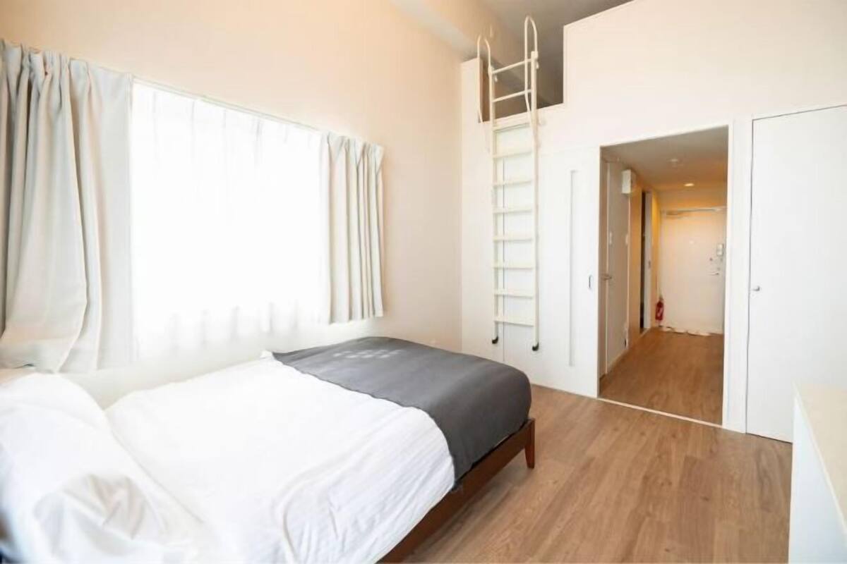Een slaapkamer met een bed, een raam met gordijnen, een houten vloer, een ladder en een deur naar een andere kamer.