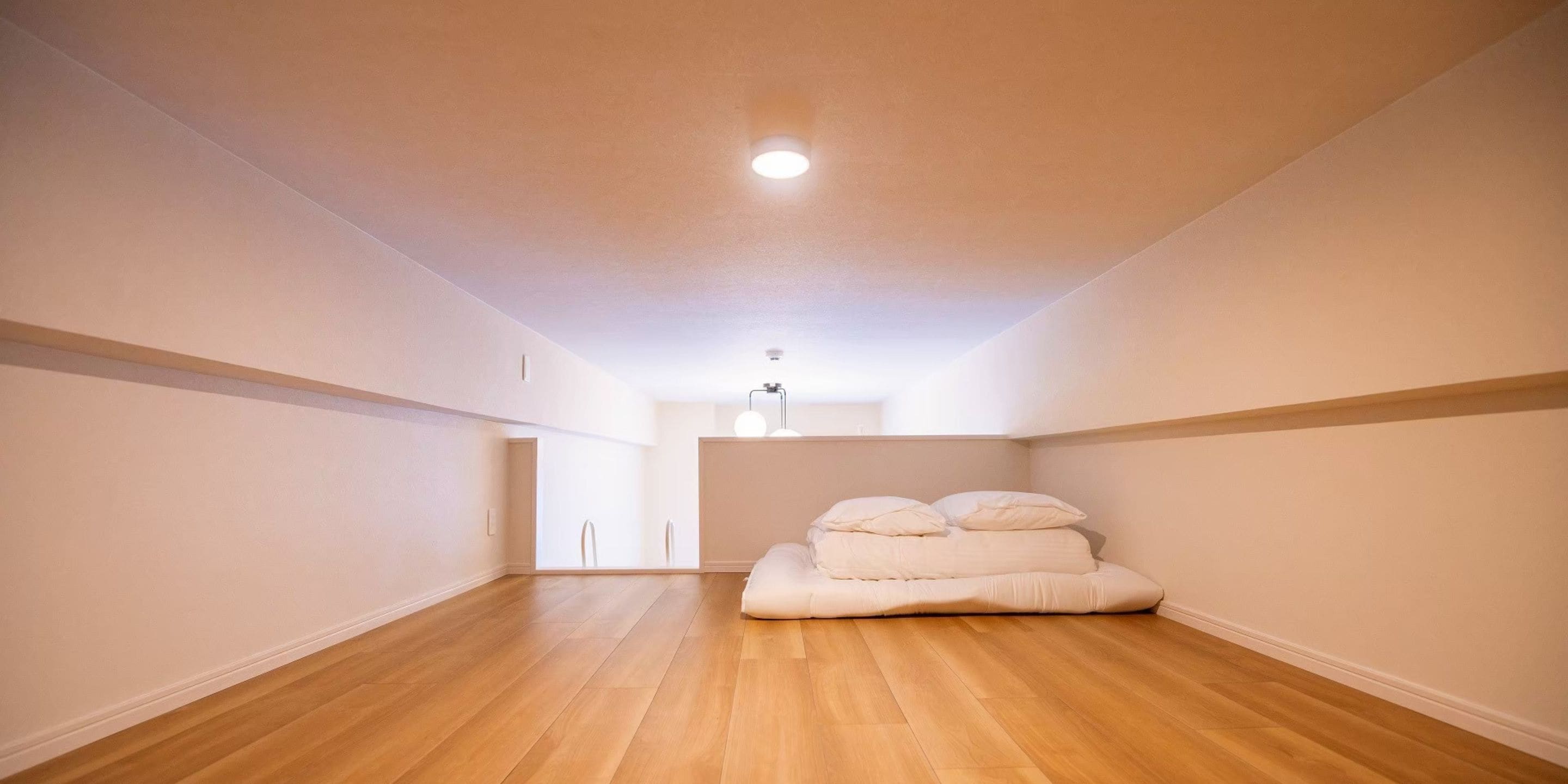 Une pièce avec un plancher en bois, un lit avec des oreillers, un luminaire au plafond et un mur blanc.