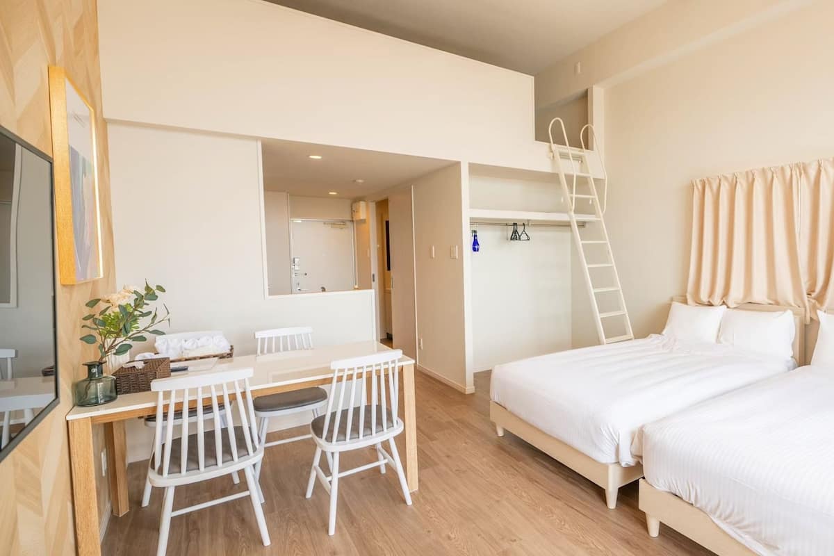 Een slaapkamer met twee bedden, een bureau met stoelen, een ladder en een zichtbare badkamer door een open deur.