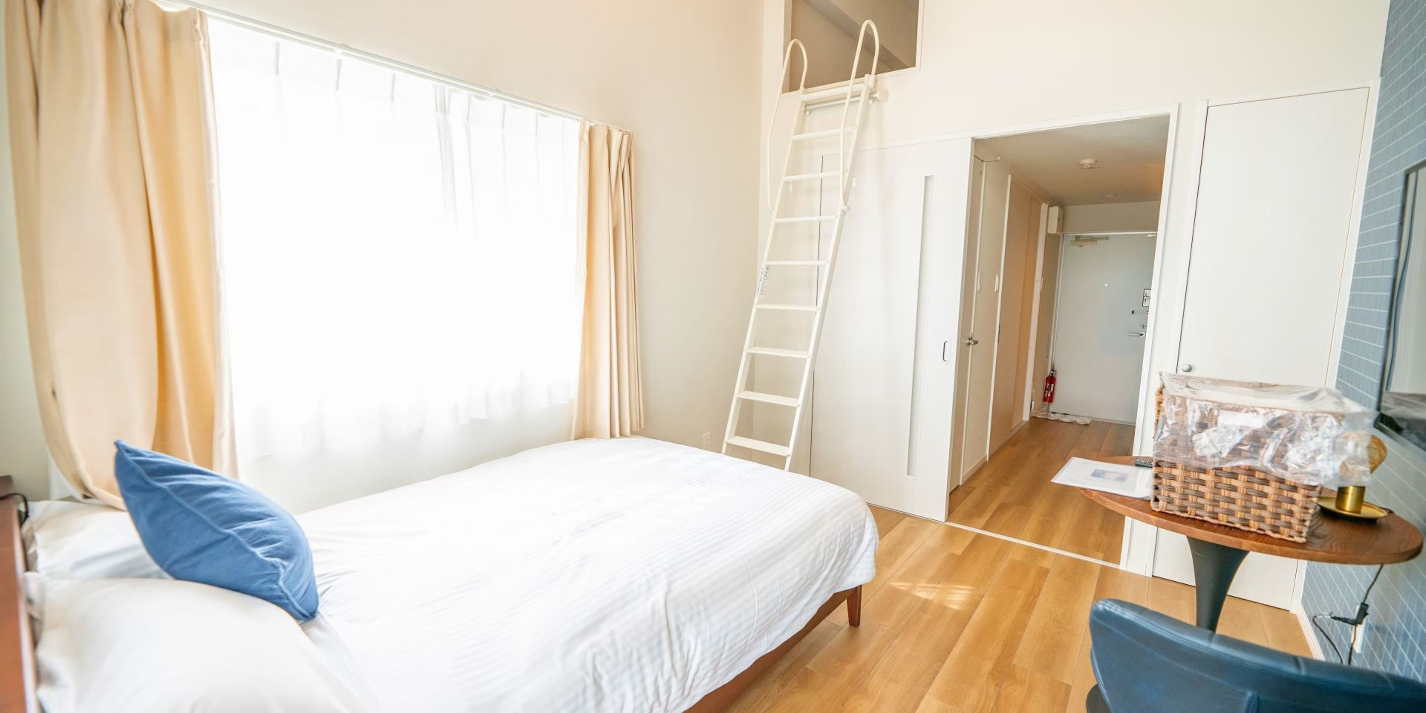 A bedroom with a bed, a ladder, a small table with a basket, and a bathroom visible through an open door.