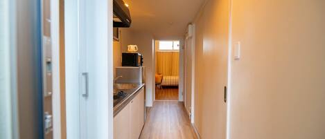 A narrow hallway with a kitchenette on the left, a bed with a striped blanket in the middle, and a window with curtains on the right.