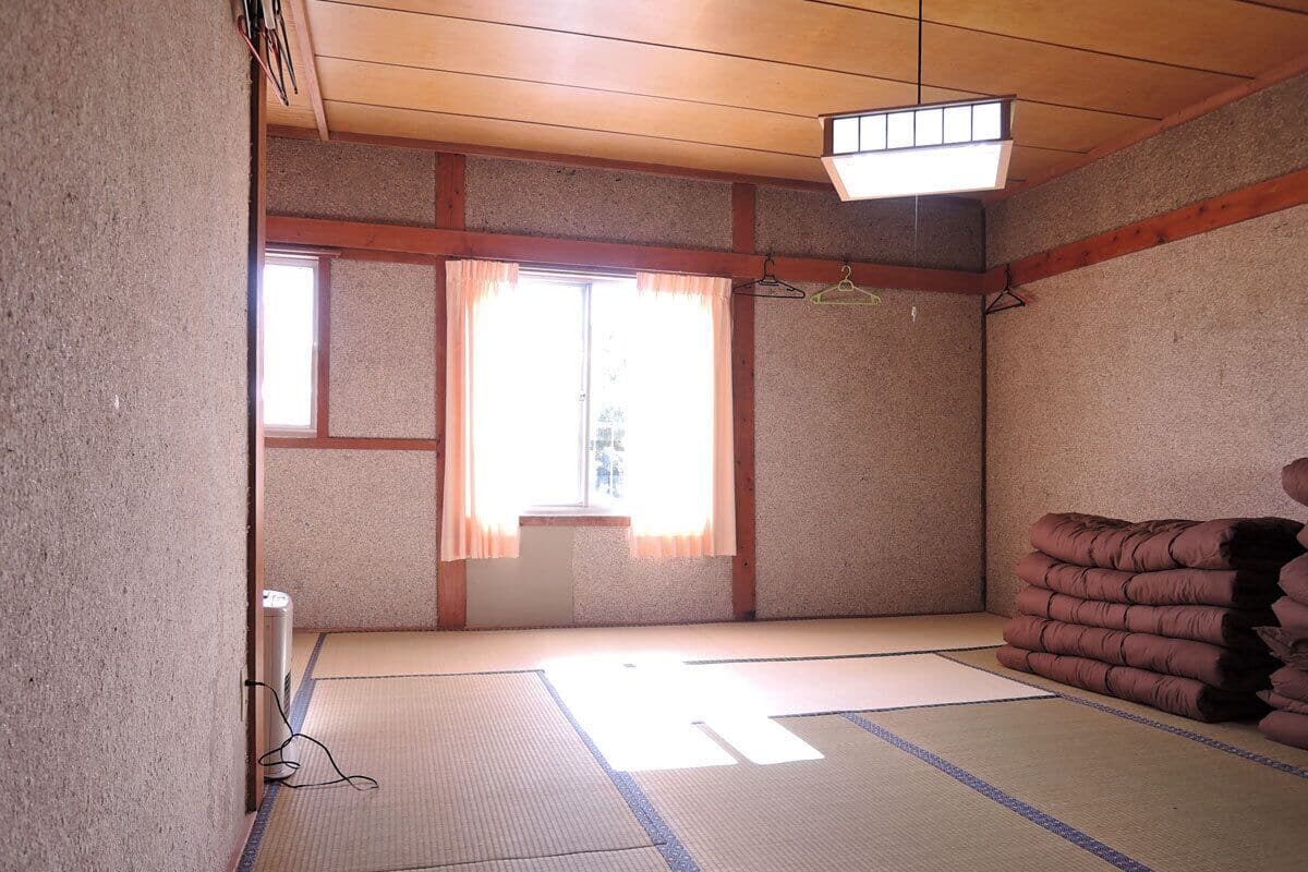 Een kamer met tatami matten, een houten plafond, een raam met gordijnen en een stap kussens.