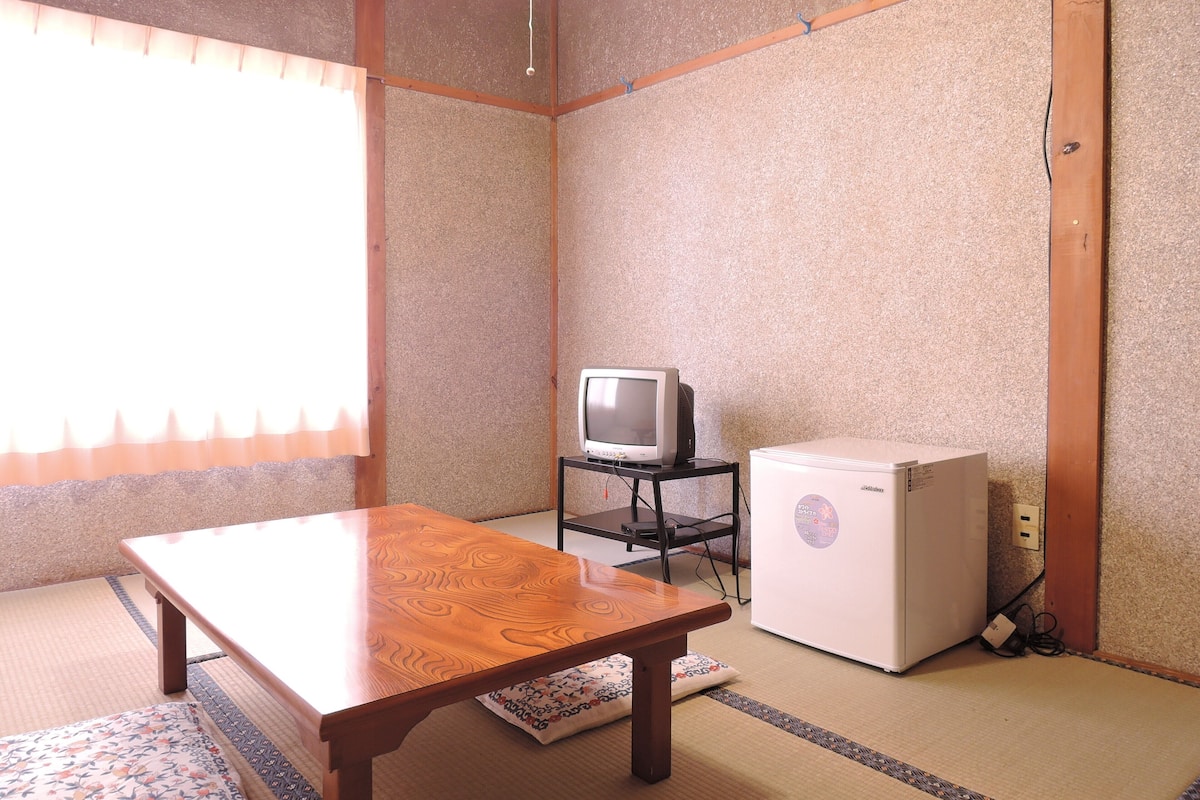 Een traditionele Japanse kamer met tatami vloerbedekking, een houten tafel, een televisie op een standaard en een koelkast.