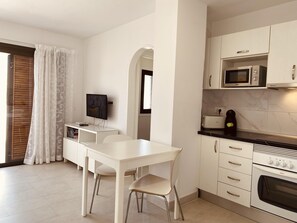 Una cocina moderna con armarios blancos, un microondas, un tostador y un horno. Hay una mesa de comedor con dos sillas y un televisor de pantalla plana sobre un mueble.