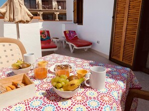 Una mesa servida con frutas, bebidas y un bol de aperitivos sobre un mantel estampado, con un balcón y sillones de descanso al fondo.