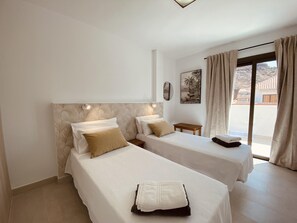 A hotel room with two single beds, a small wooden table, a window with curtains, and a view of buildings outside.
