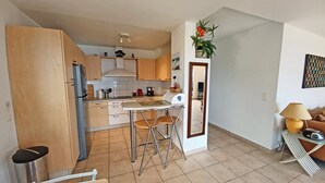 A compact kitchen with a refrigerator, sink, and bar stools. There is a dining area with a table and chairs, and a small seating area with a lamp. - T3 of 68 m2 in Porticcio. Covered terrace 20 m2, small garden 50 m2. Walking distance to the beach. (Grosseto-Prugna)