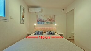 A bedroom with a bed measuring 160 cm, a cityscape painting above the bed, an air conditioner on the wall, and a bedside table with a lamp. - T3 of 68 m2 in Porticcio. Covered terrace 20 m2, small garden 50 m2. Walking distance to the beach. (Grosseto-Prugna)