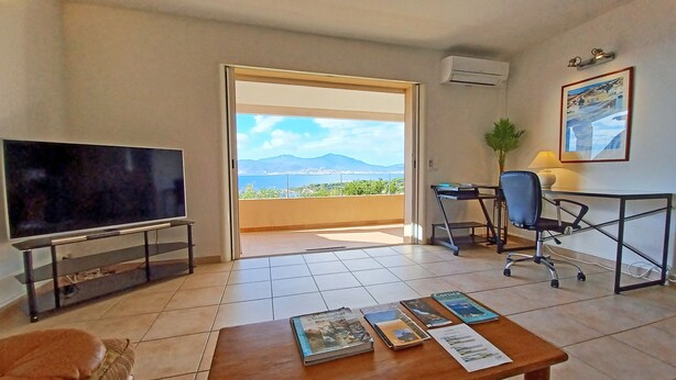 A room with a TV, a desk with a chair, a table with books, and a view of mountains through a door. - T3 of 68 m2 in Porticcio. Covered terrace 20 m2, small garden 50 m2. Walking distance to the beach. (Grosseto-Prugna)
