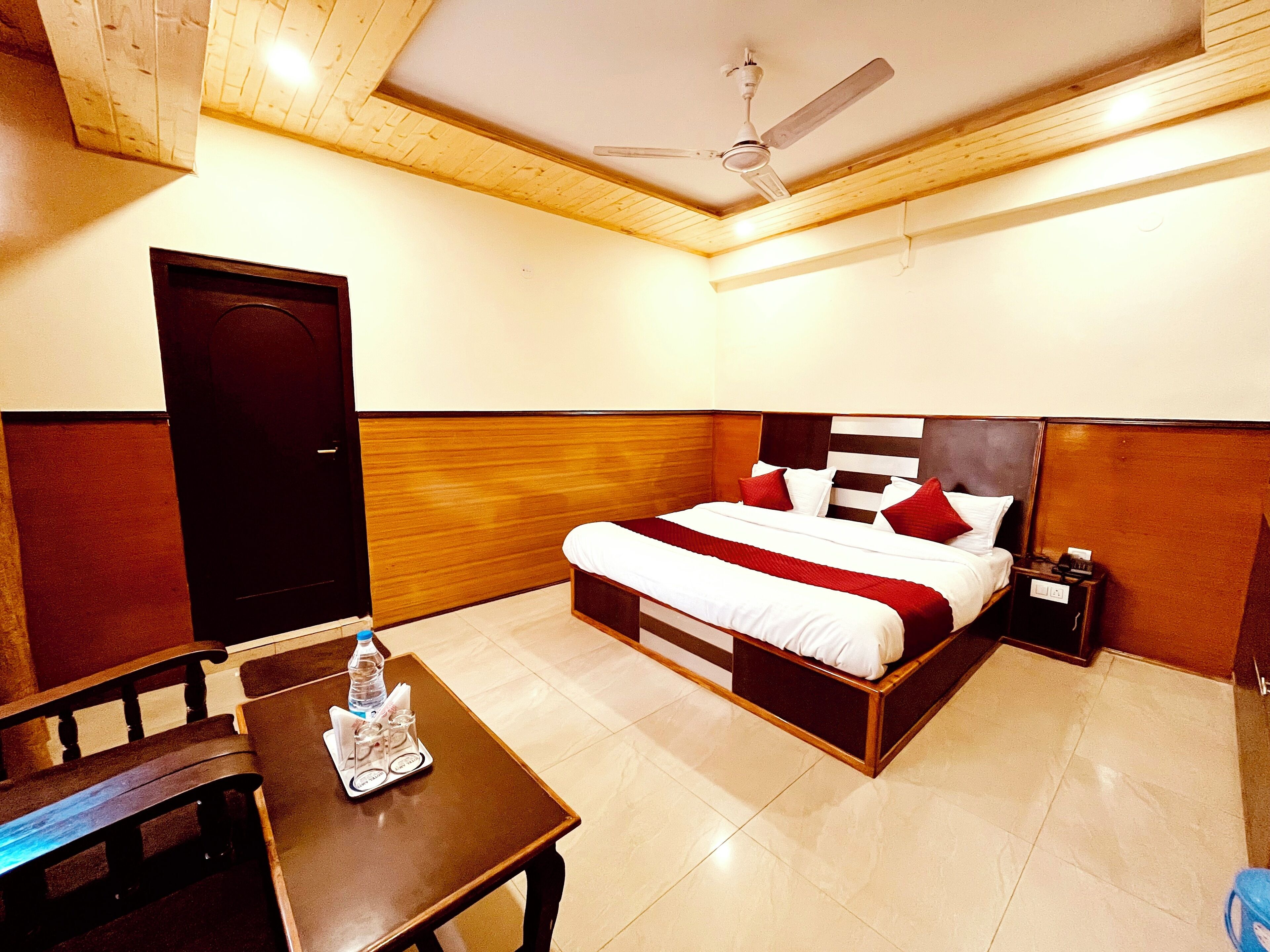 A hotel room with a bed, a wooden headboard, a nightstand, a table with a bottle and glasses, and a ceiling fan.