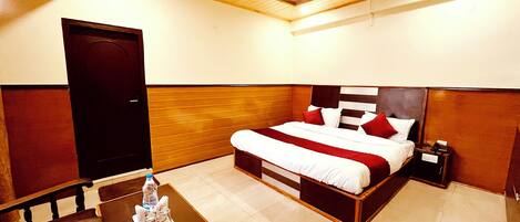 A hotel room with a bed, a wooden headboard, a nightstand, a table with a bottle and glasses, and a ceiling fan.