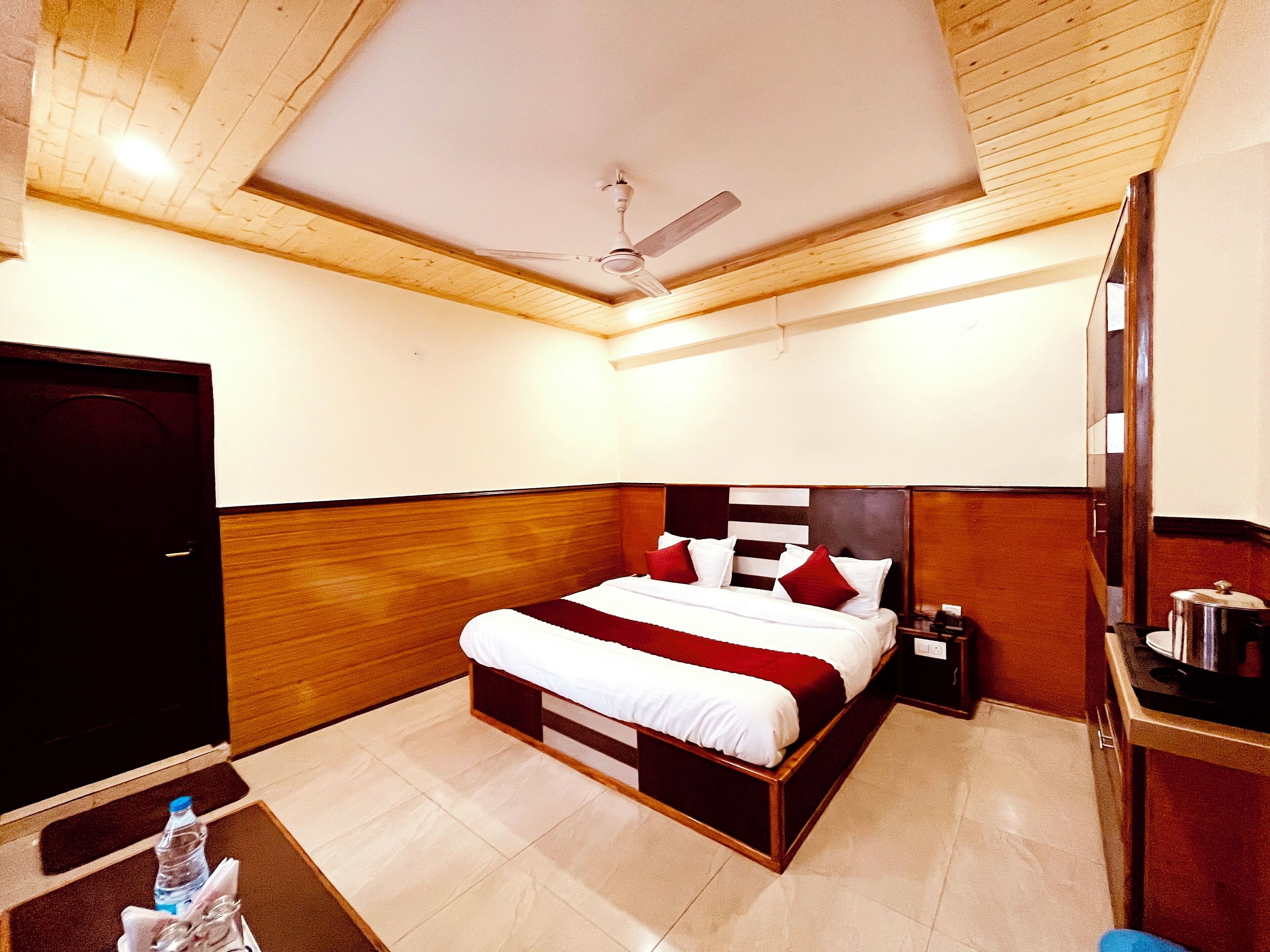 A hotel room with a wooden ceiling, a bed with white and red bedding, a nightstand, a desk with a kettle, and a water bottle on the floor.
