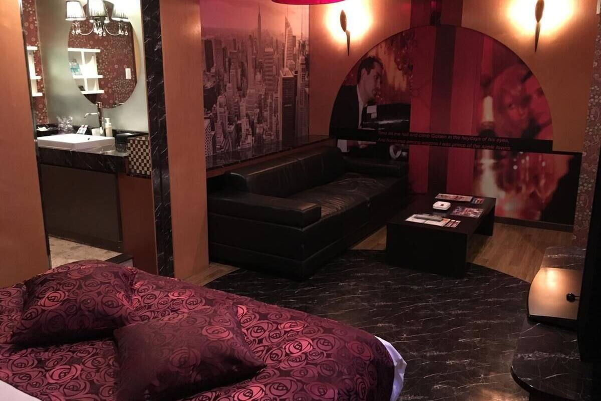Quarto de hotel moderno com uma cama grande, área de estar com sofá, uma mesinha e um mural com vista da cidade na parede.