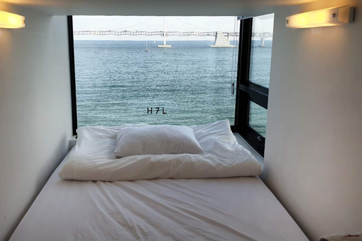 A small, single-bed room with a view of a bridge and the sea through large windows.
