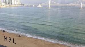 A beach with the letters "H7L" on the sand, a bridge in the background, and a city skyline across the water.