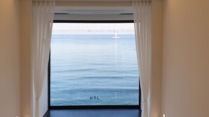 A window with white curtains, a view of the sea, and a bridge in the distance.