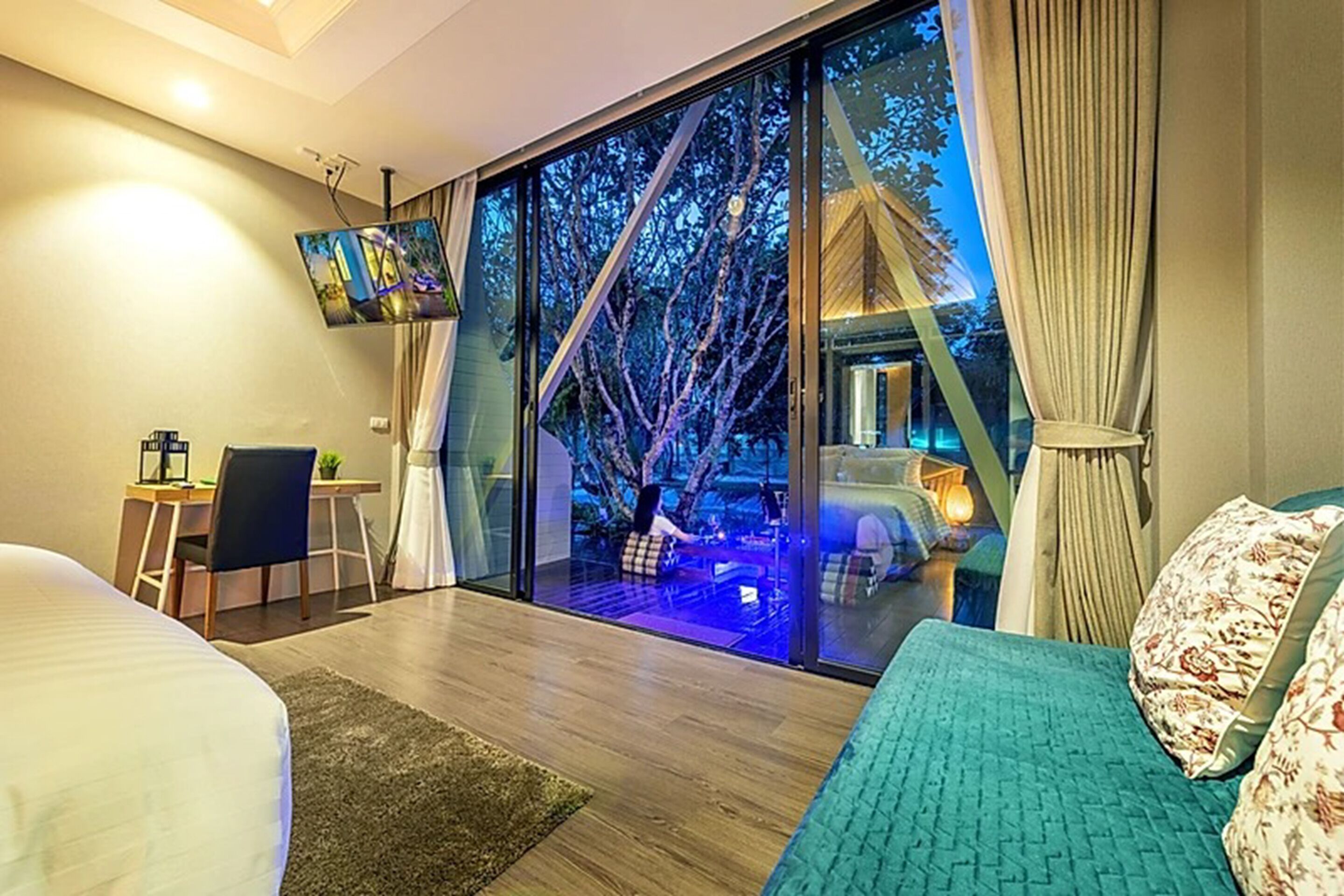 A modern hotel room with a large sliding glass door leading to an outdoor area. There is a bed with a blue bedspread, a desk with a chair, and a flat-screen TV mounted on the wall.