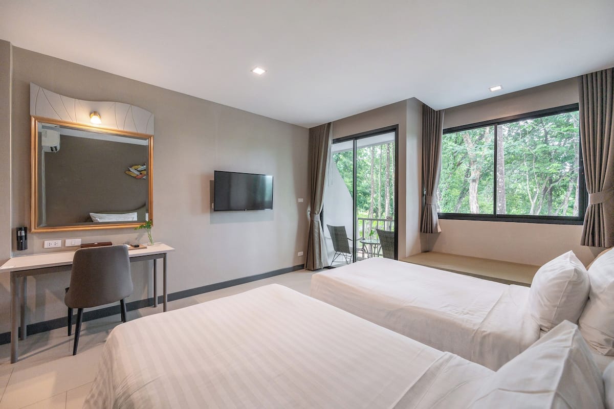 A hotel room with two beds, a desk with a chair, a TV, and a balcony with a view of trees.