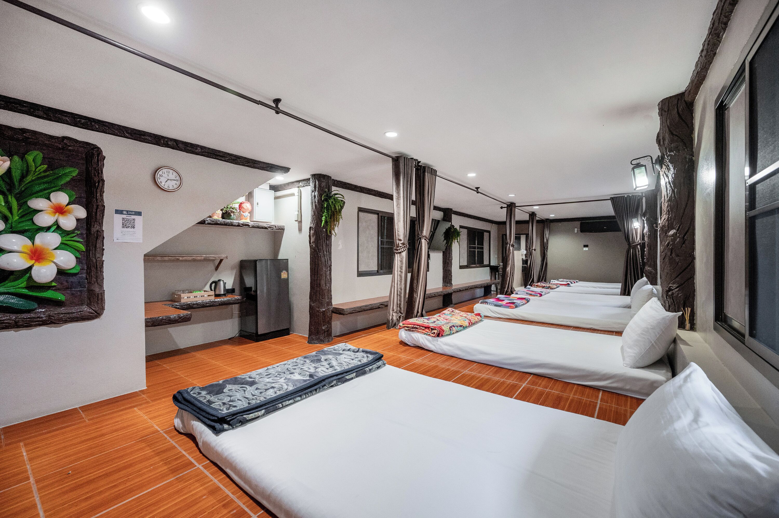 Una habitació d'hotel amb tres llits individuals, una cuineta petita, un rellotge i una planta decorativa.
