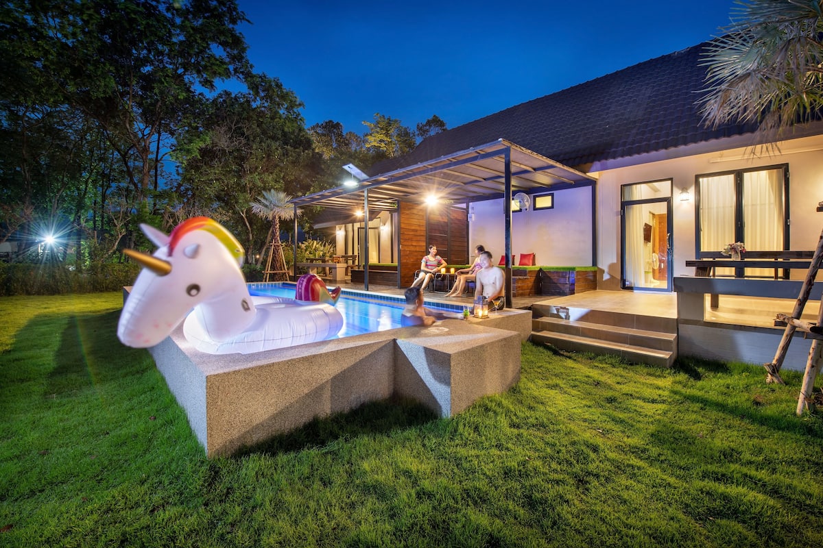 A swimming pool with a unicorn float, a covered patio, and a house with a wooden deck and a door.