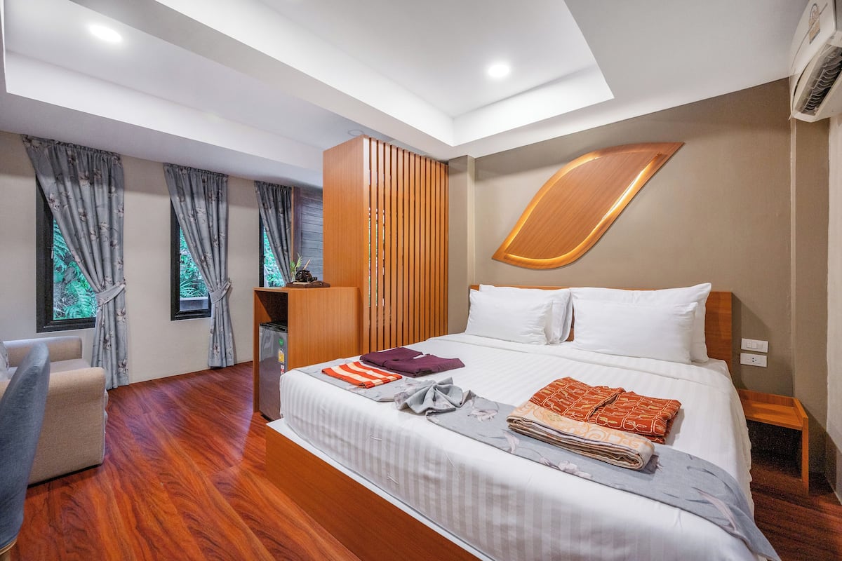 A modern hotel room with a large bed, a wooden wardrobe, a desk, and a view of greenery through the windows.