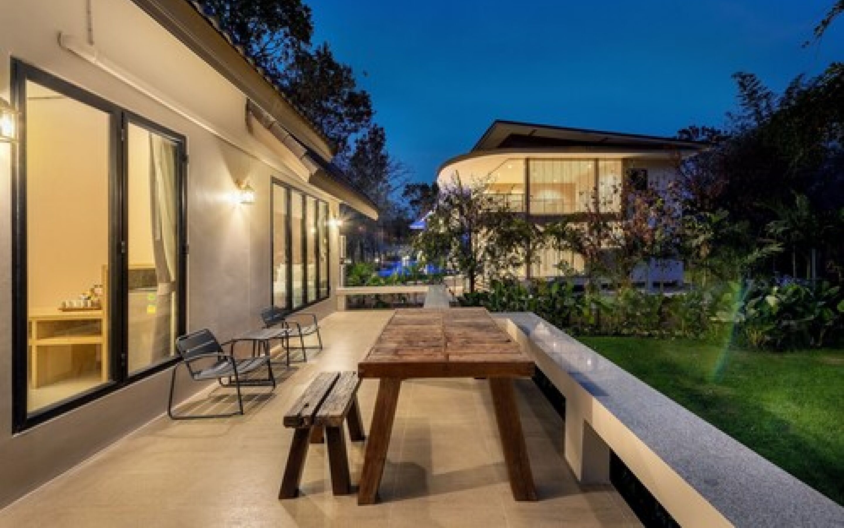 Un patio exterior moderno con una mesa y bancos de madera, rodeado de un jardín cuidado y un edificio con grandes ventanales.