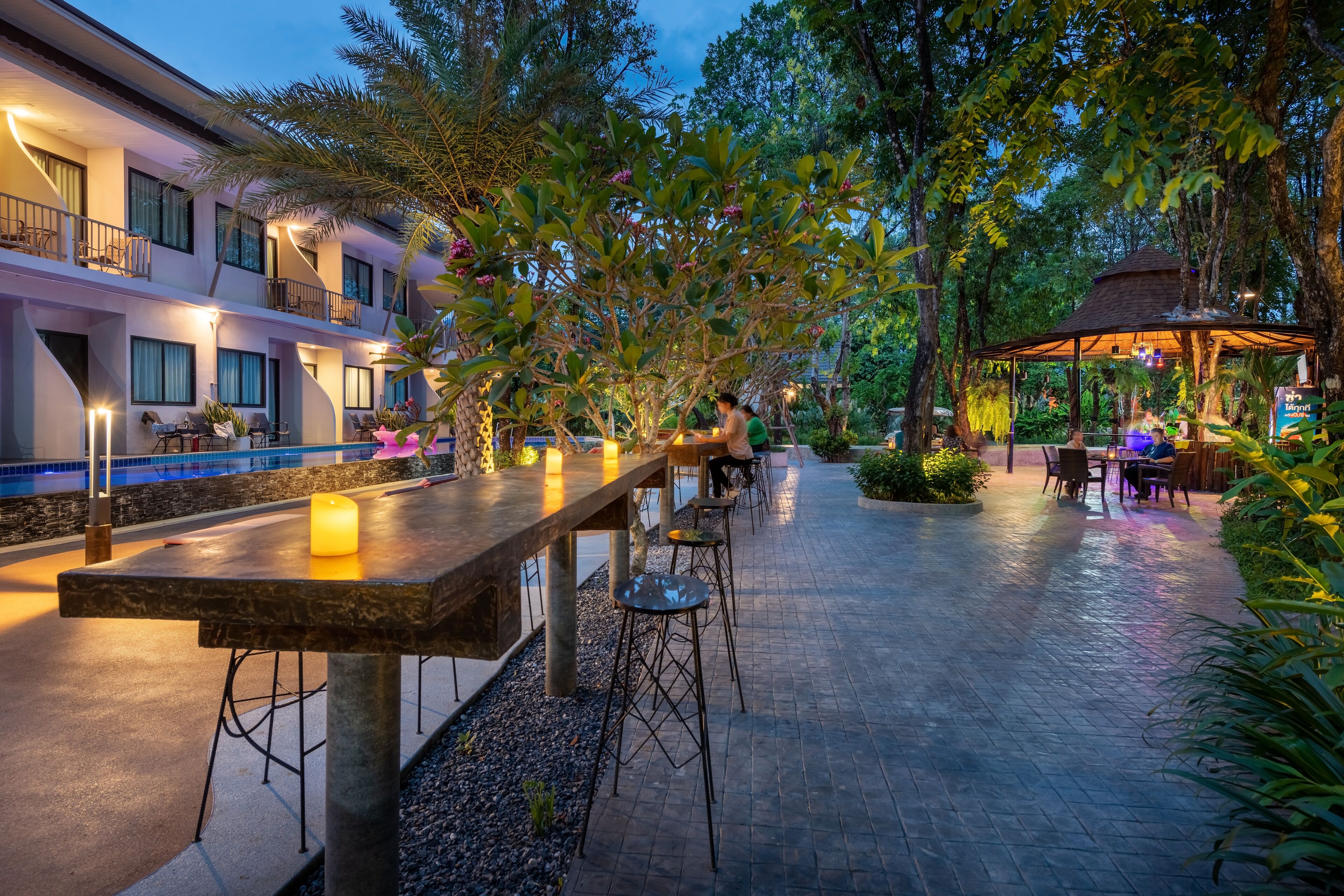 An outdoor patio area with a long wooden table, bar stools, and a pool in the background.