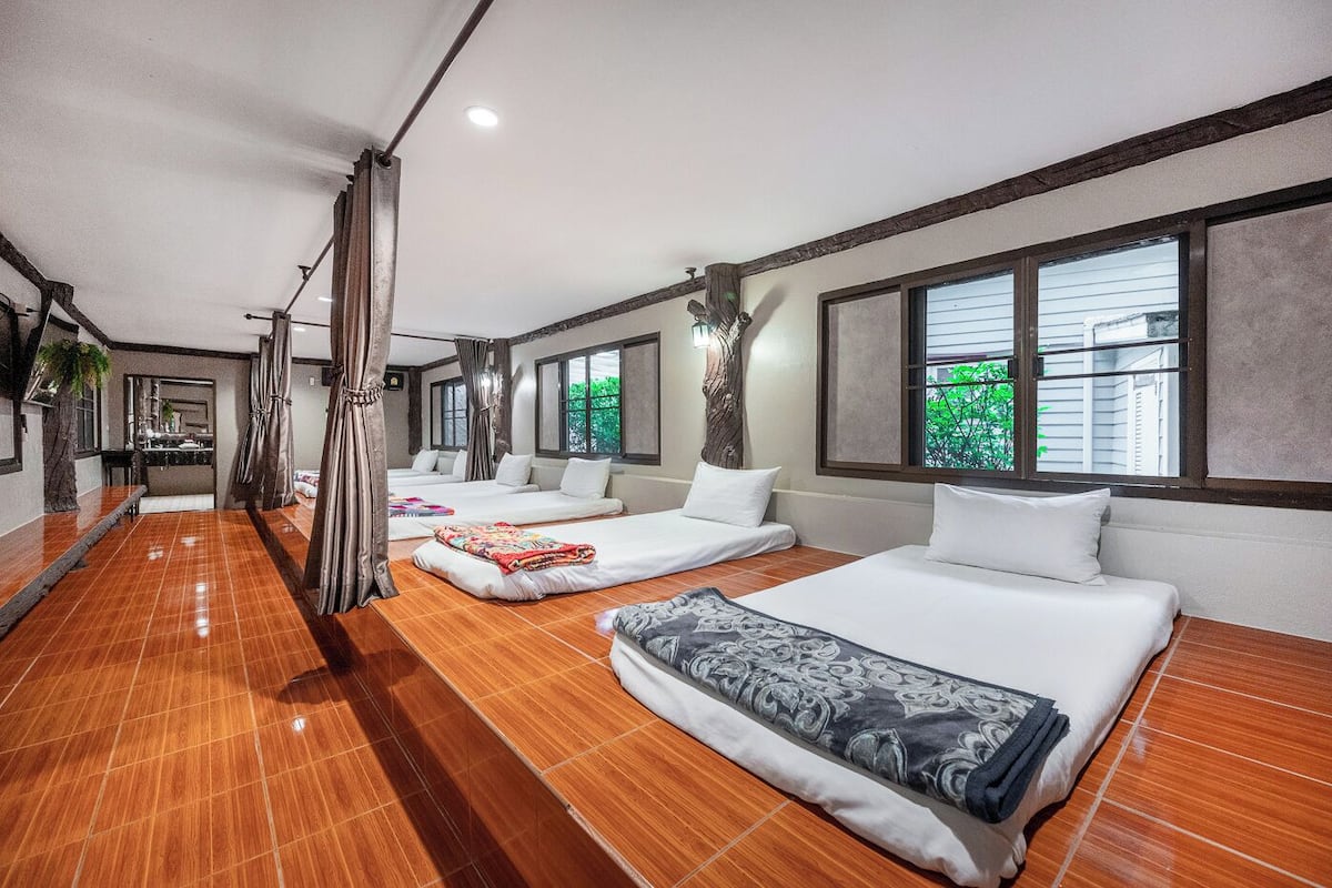 A hotel room with three beds, a wooden floor, large windows, and a tree outside.