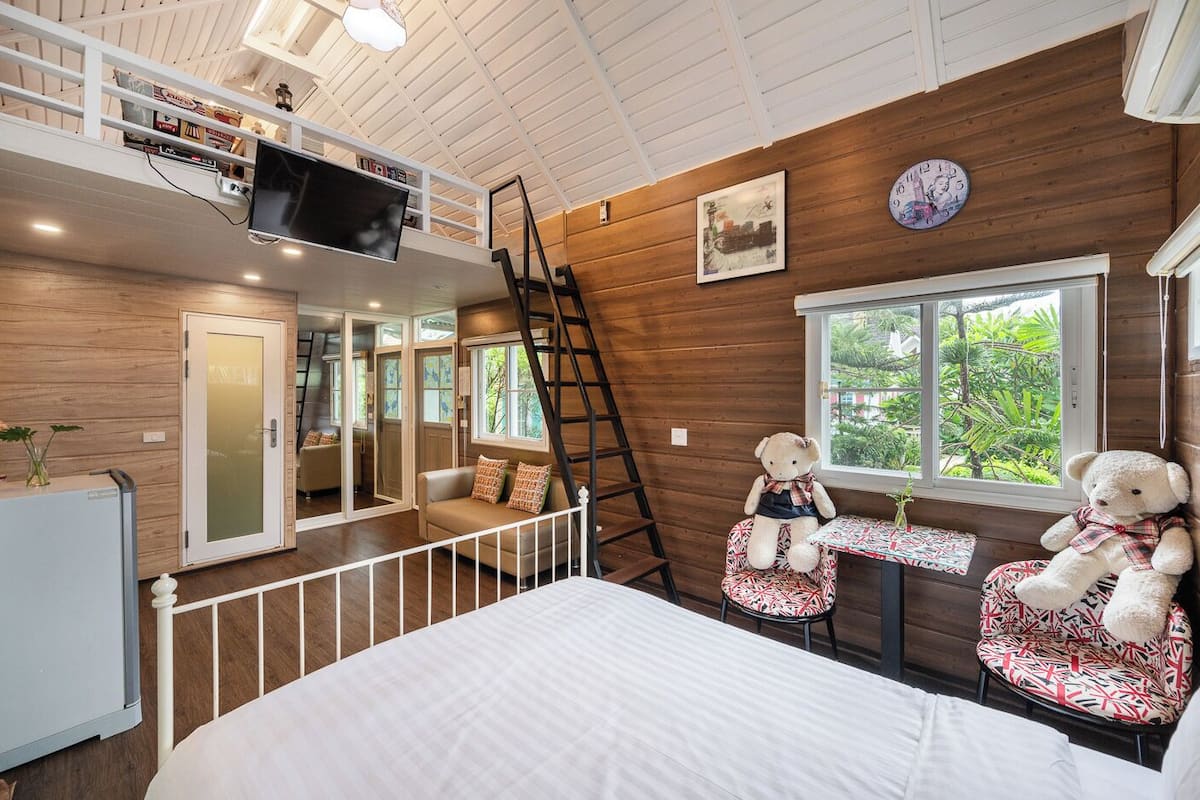 A cozy room with a bed, a desk with a teddy bear, a TV on the wall, and a window with a view of greenery.