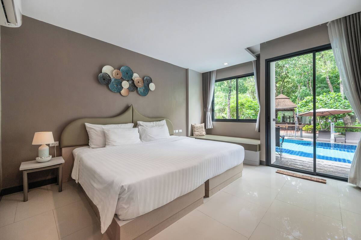 A modern hotel room with a large bed, a bedside table with a lamp, a decorative wall piece, and a view of an outdoor swimming pool.
