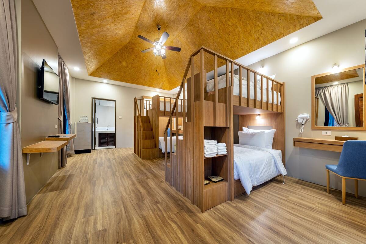 A modern hotel room with a wooden ceiling, bunk beds, a desk, a chair, a mirror, and a bathroom with a shower.