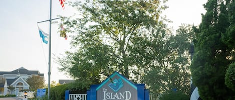 A sign for 'The Island at Hidden Harbour' with flags, a tree, and a building in the background.