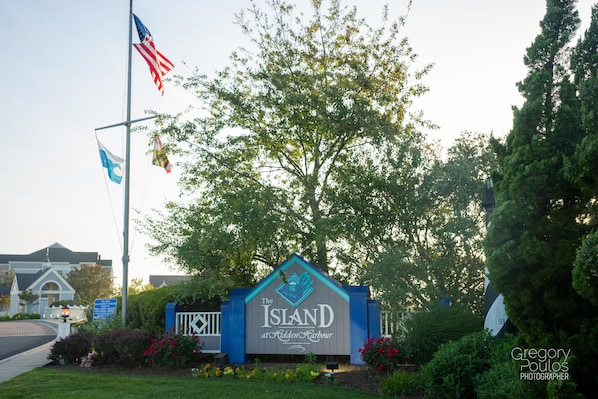 A sign for 'The Island at Hidden Harbour' with flags, a tree, and a building in the background. - Sunsets, Peace & Quiet, Next to Northside Park, Over 60 5 Star Reviews! (Ocean City)