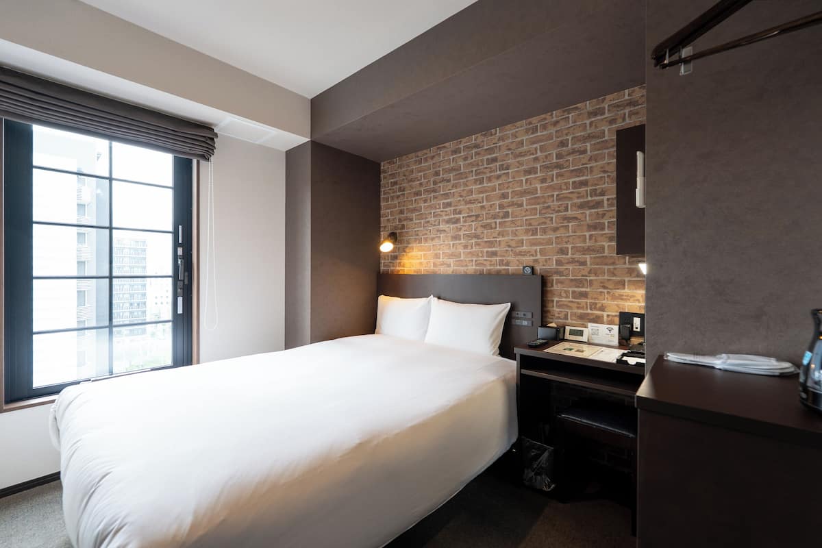 Hotelkamer met een groot bed, een wand van baksteen, een bureau met computer en een raam met jaloezieën.