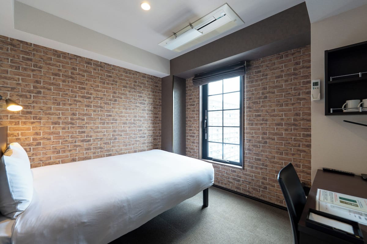 Hotelkamer met een bakstenen muur, een enkel bed, een bureau met stoel, een raam met zonwering en een thermostaat aan de muur.