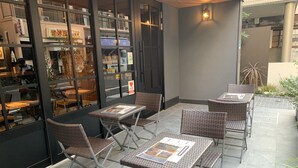 An outdoor seating area with wicker tables and chairs, a tiled floor, and a glass wall with reflections of the interior. - Hotel Tabard Tokyo (Tokyo)