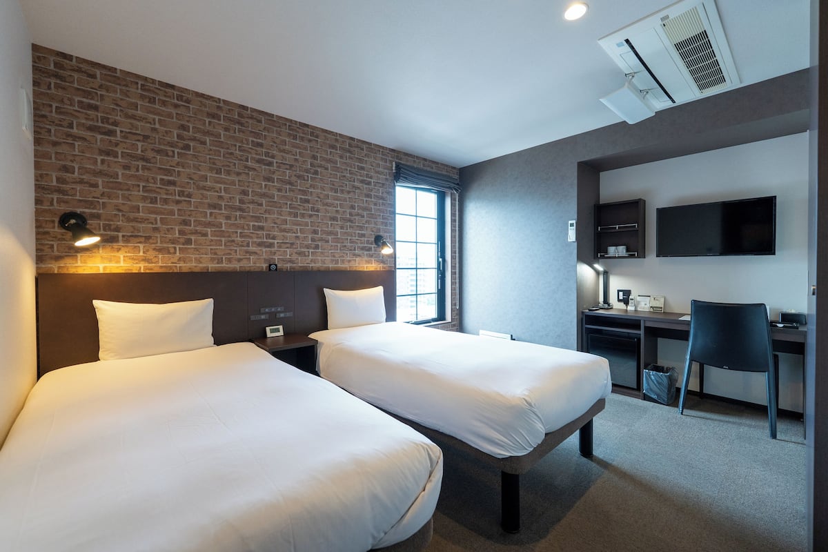 Een hotelkamer met twee bedden, een bureau, een stoel, een televisie en een raam met uitzicht op de stad.