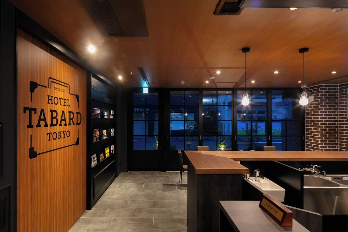 Een moderne hotelentree met een houten wand waarop 'HOTEL TABARD TOKYO' staat, een receptiebalie, een bar en grote ramen met uitzicht over de stad.