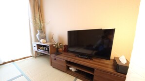 A room with a wooden TV stand, a flat-screen TV, a small table with a vase and a teddy bear, and a tatami mat floor. - EX Tenjinnomori Apartment 202 (Osaka)