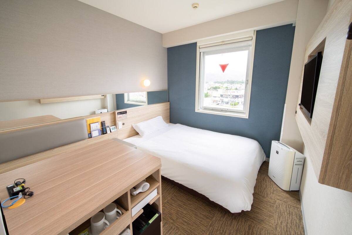 A hotel room with a bed, bedside table, and a small desk with a chair. There is a window with a view of buildings outside.