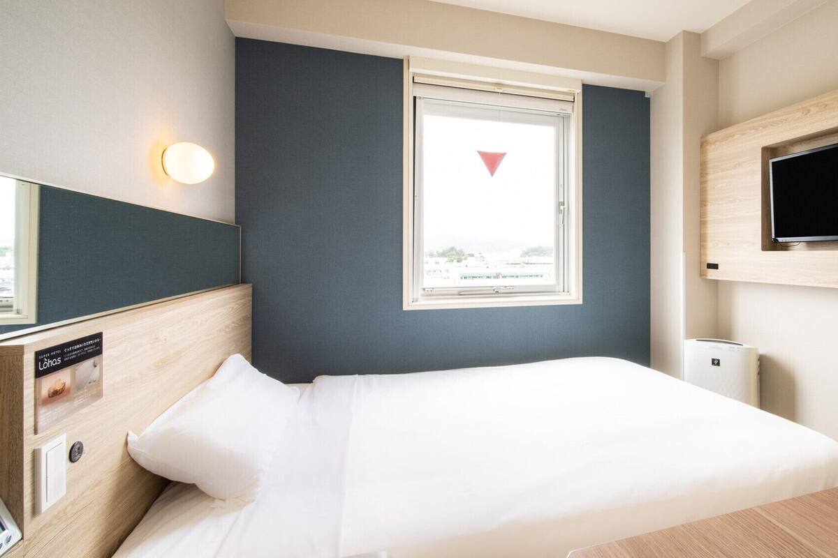 A hotel room with a bed, a TV mounted on the wall, a window with a view, and a thermostat on the wall.