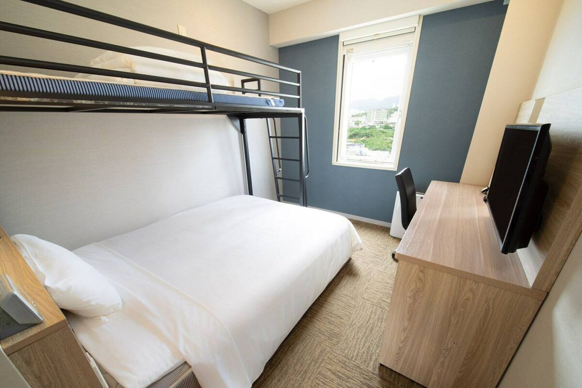 A hotel room with a bunk bed, a desk with a flat-screen TV, and a window with a city view.