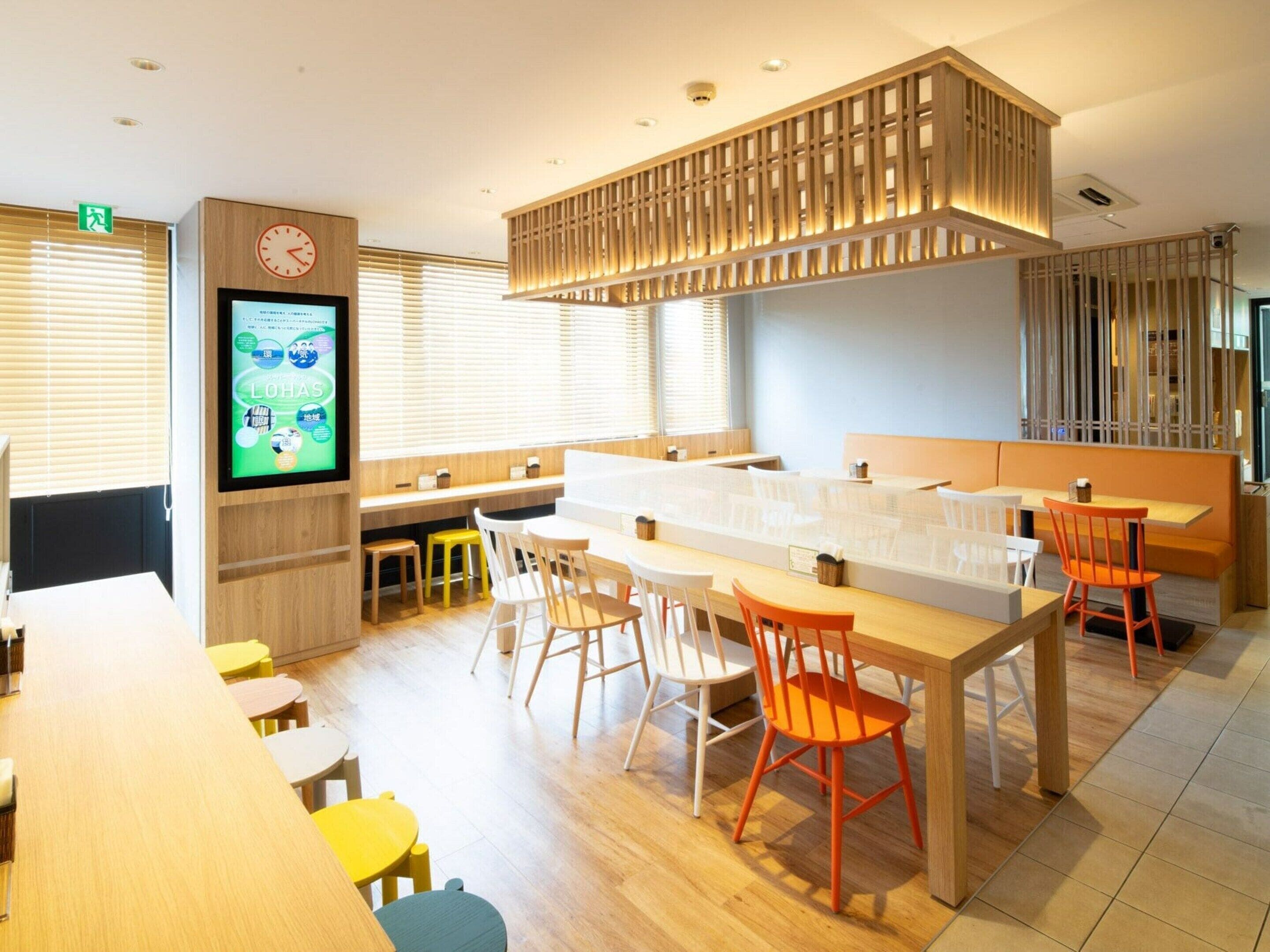 Un restaurante moderno con mobiliario de madera, una pantalla digital para el menú y un área de estar con techo alto y listones de madera decorativos.