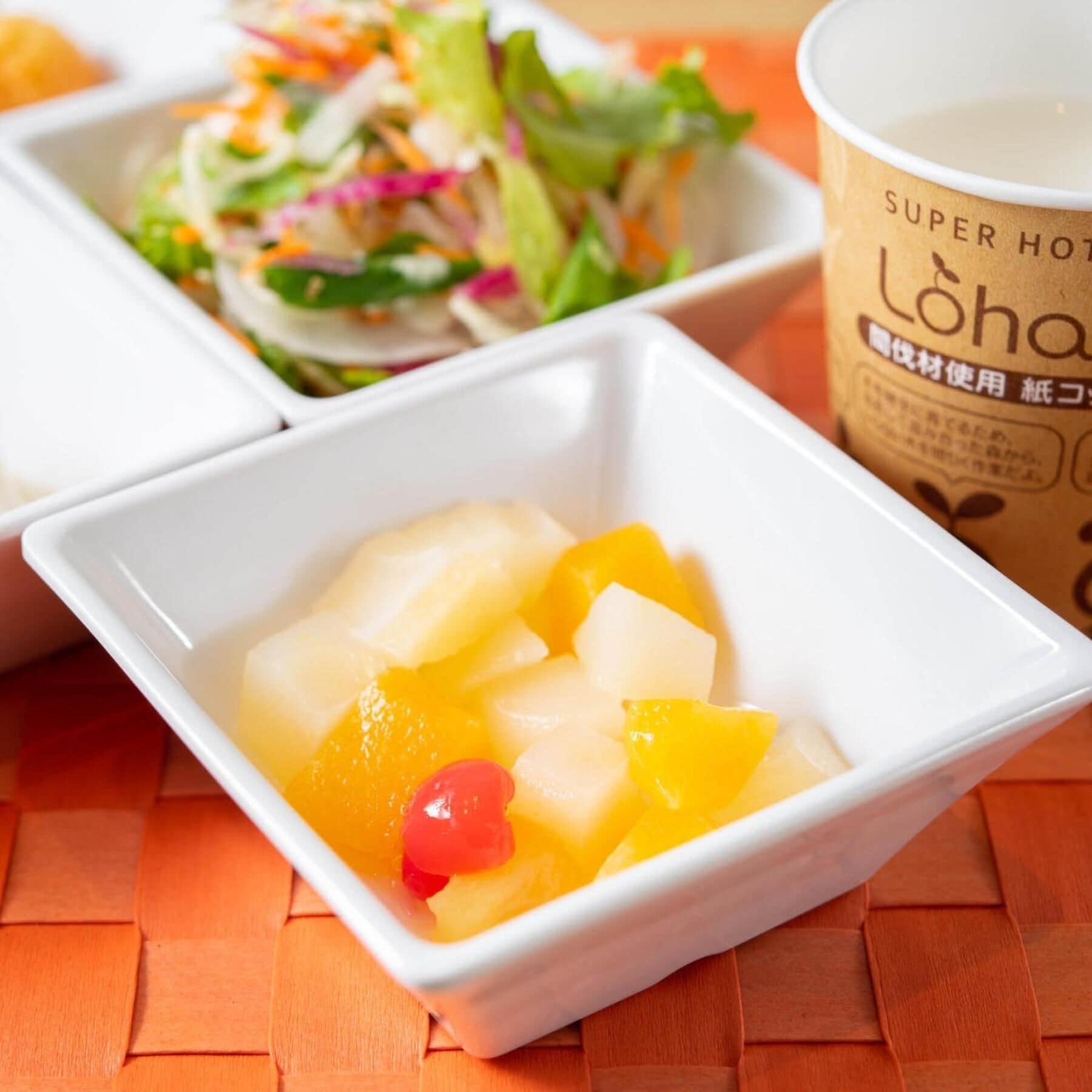 Eine weiße Schüssel mit gewürfeltem Obst und einer roten Kirsche, ein Salat in einer weißen Schüssel und eine Tasse mit dem Aufdruck 'Super Hot' auf einer orangefarbenen gewebten Unterlage.