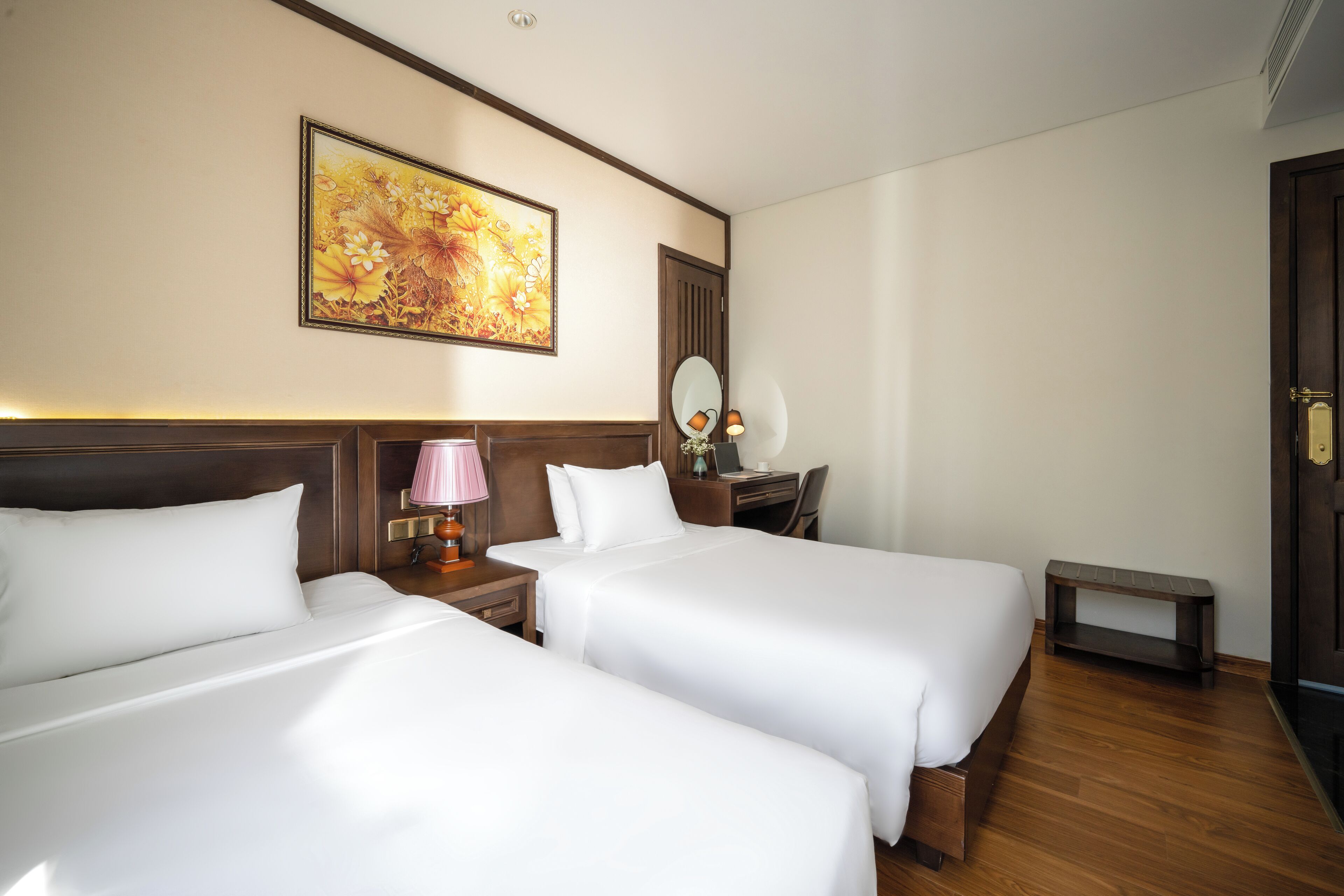 A hotel room with two beds, a desk, a chair, a small table, a lamp, and a painting on the wall.