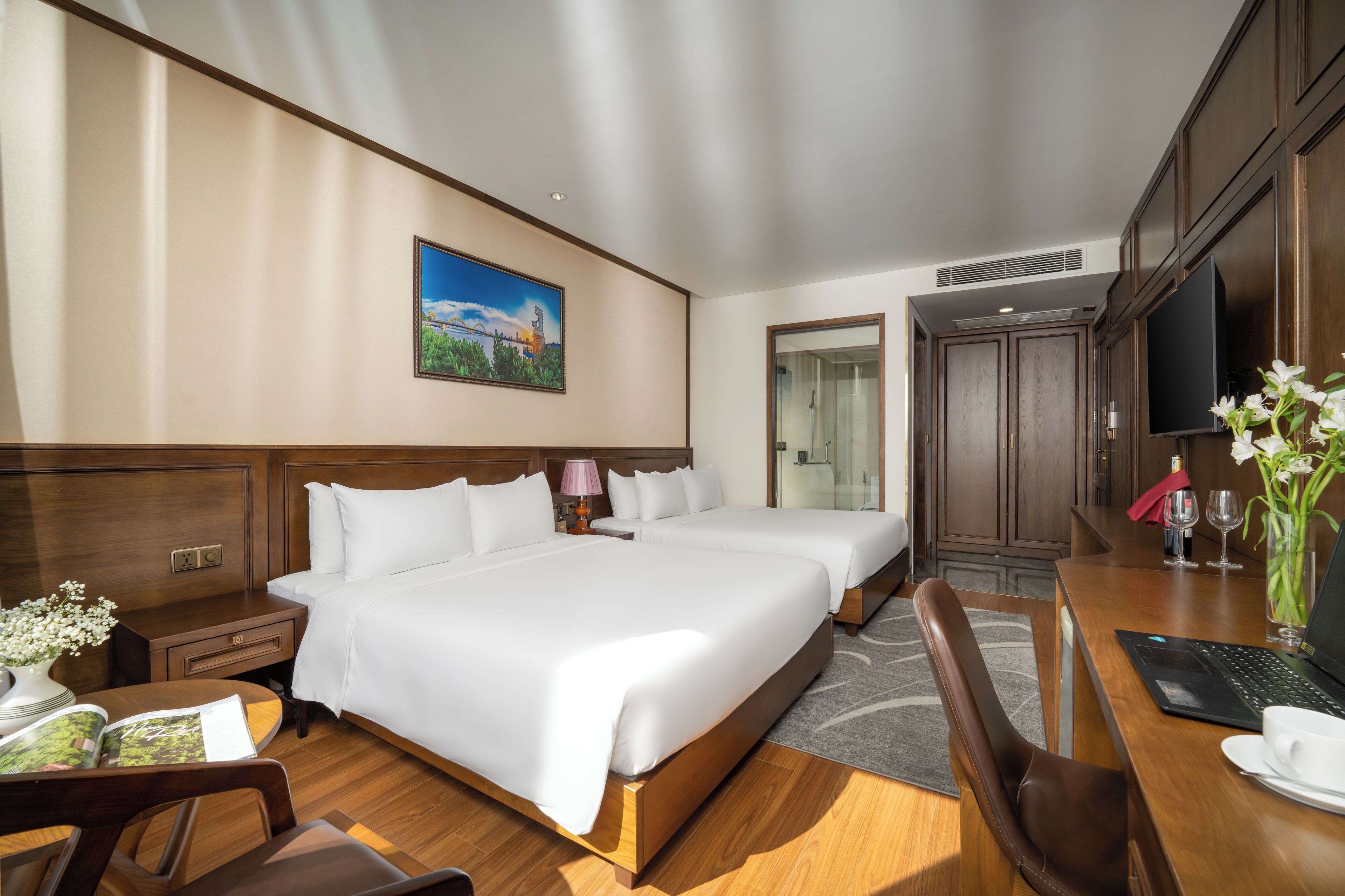 Habitación de hotel con dos camas, un escritorio con una computadora portátil, un televisor y un cuadro en la pared.