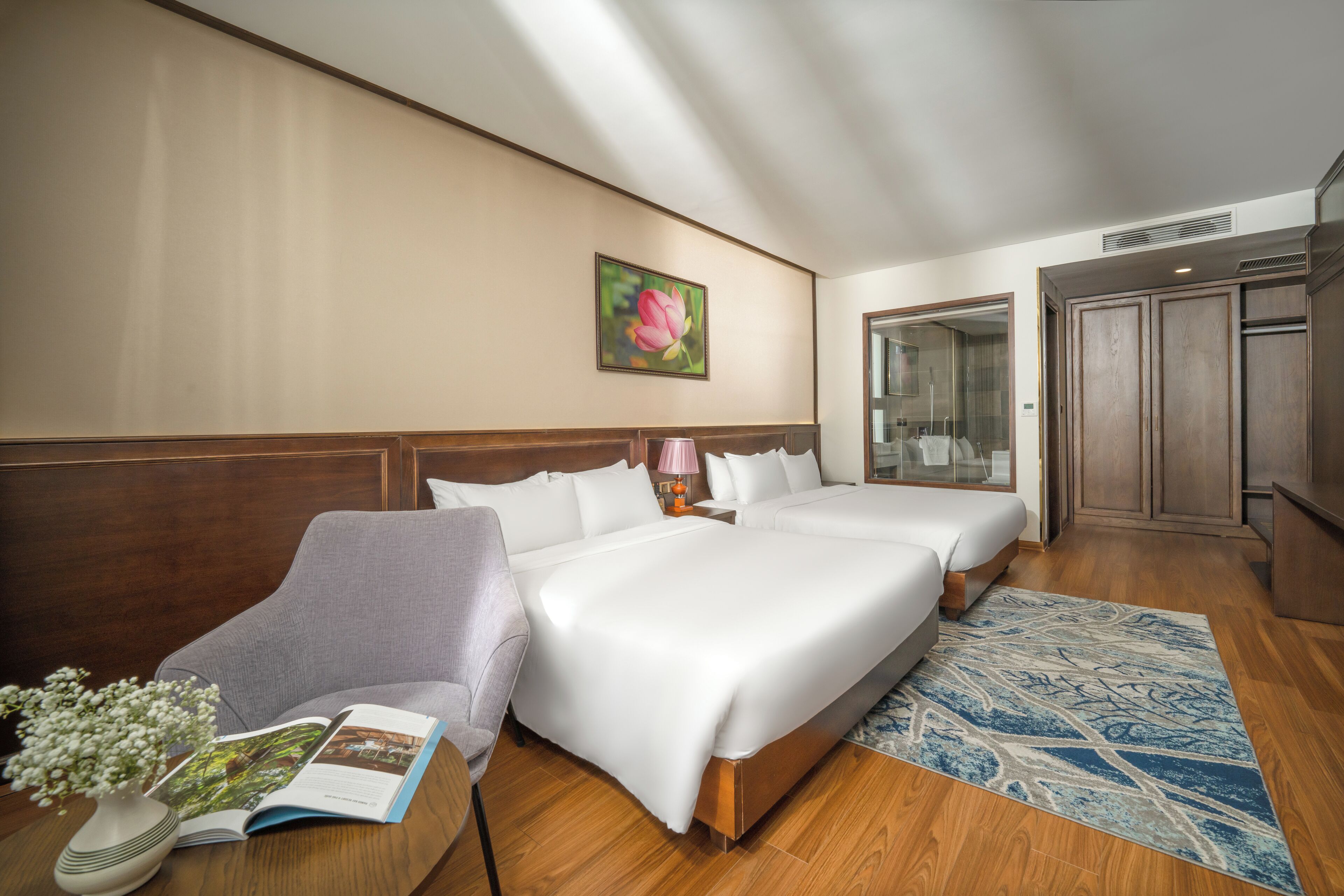 A hotel room with two beds, a sitting area with a chair, a bedside table with a lamp, a wooden floor, a large window, and a painting on the wall.