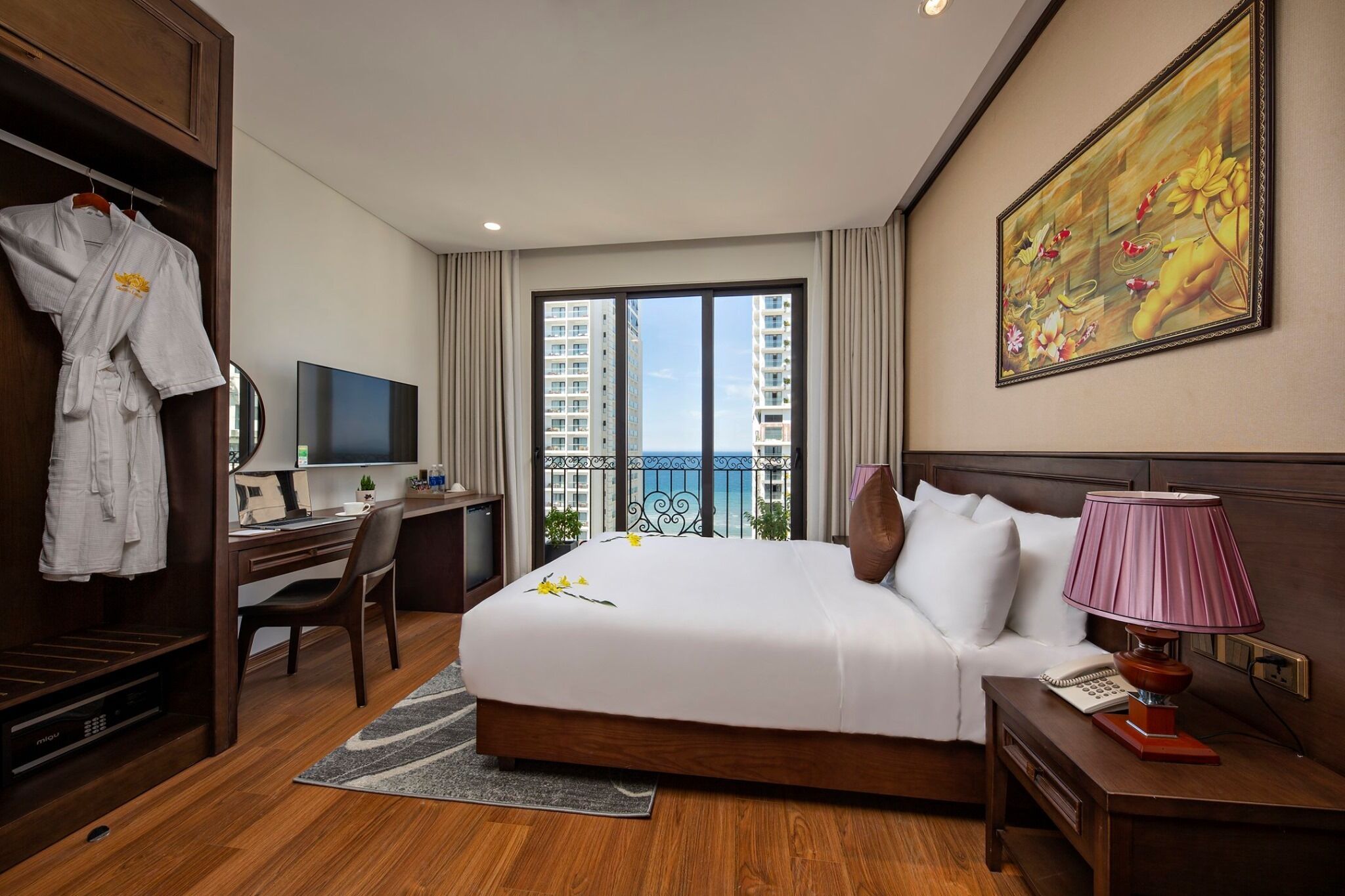 A hotel room with a bed, desk, TV, and a balcony with a view of high-rise buildings.