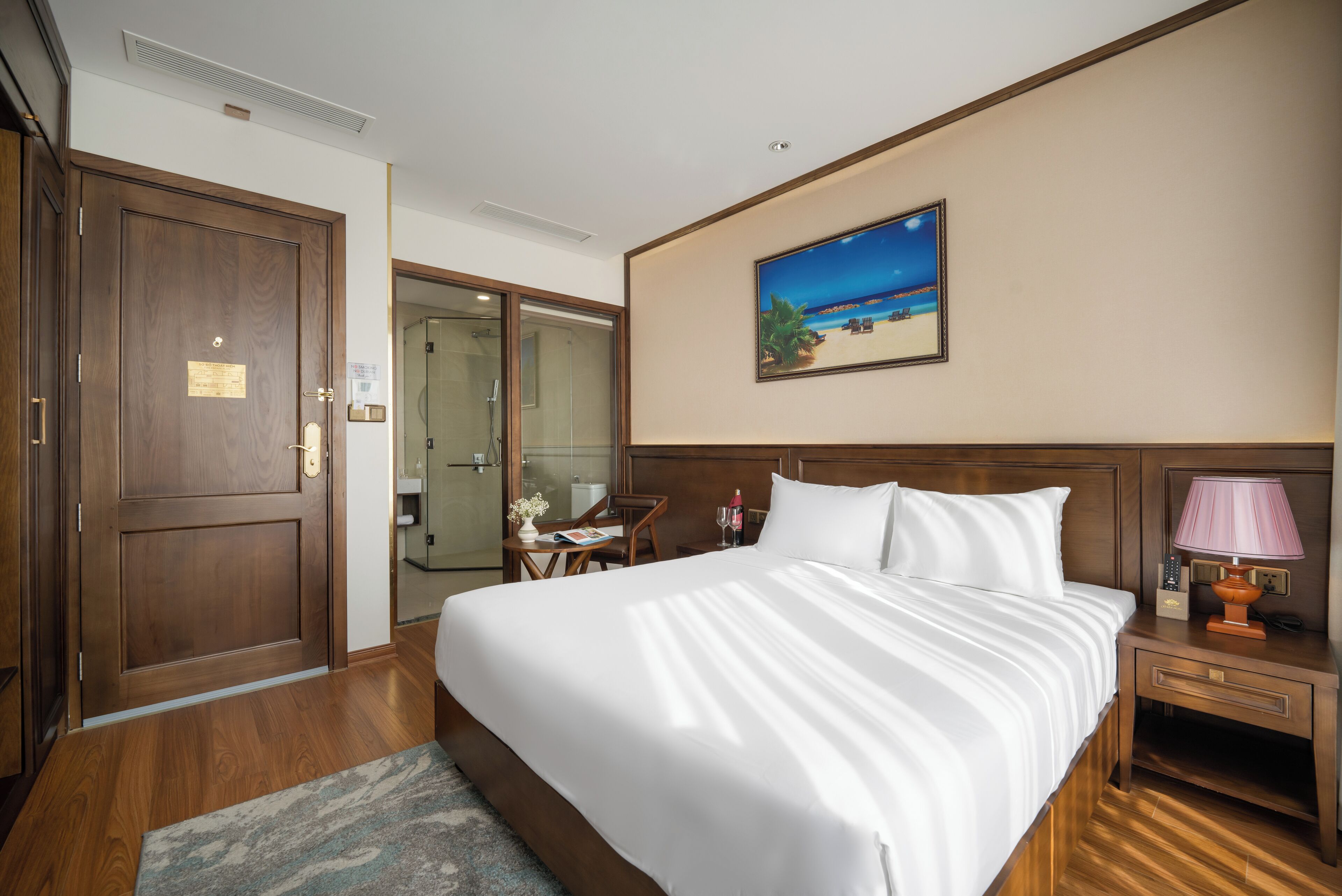 A hotel room with a large bed, a nightstand with a lamp, a painting of a beach scene, and a door leading to a bathroom.